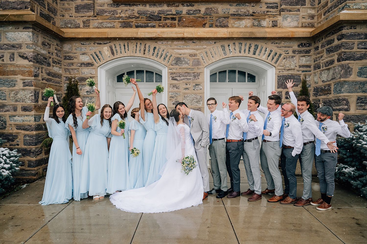 Wedding party in light blue dresses and gray suits poses together against stone building exterior.