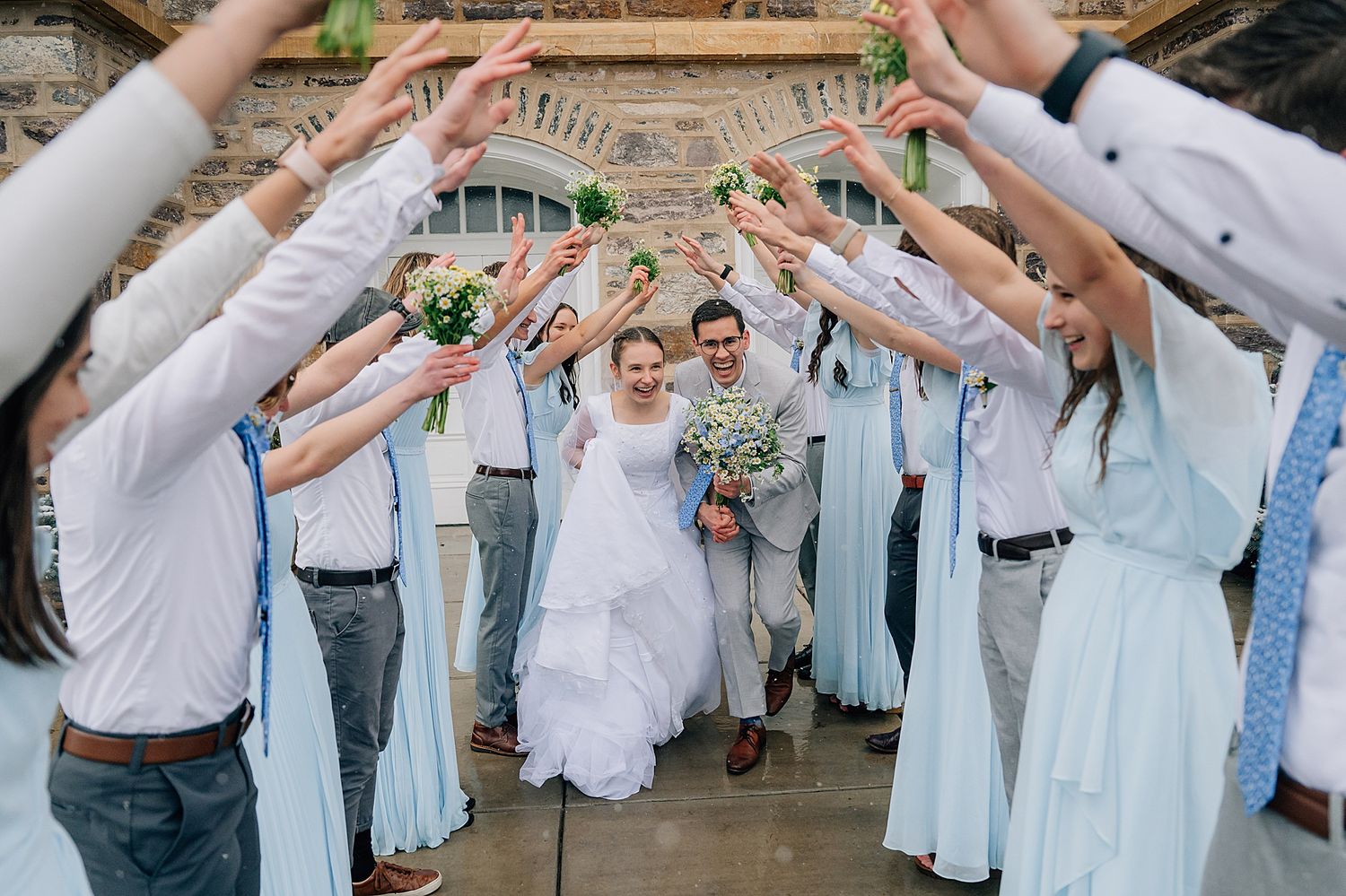 Wedding celebration with bridal party wearing light blue dresses and suspenders forming a tunnel with raised arms.