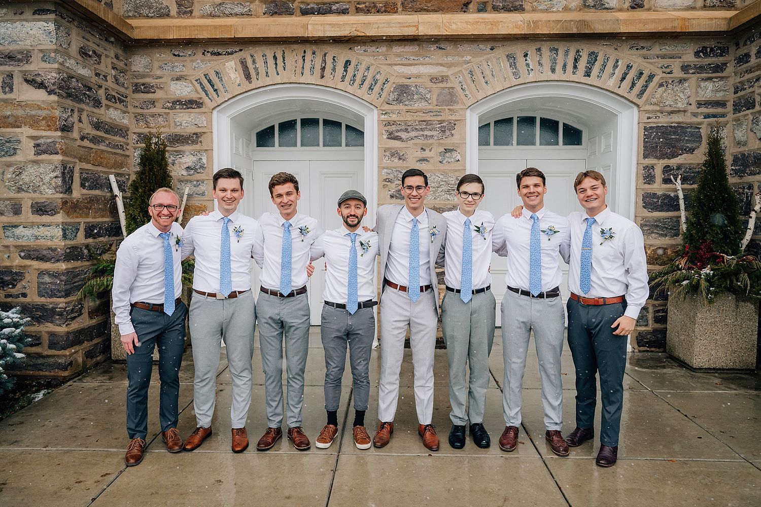 A group in matching light blue ties and gray slacks stand together against a rustic stone wall background.
