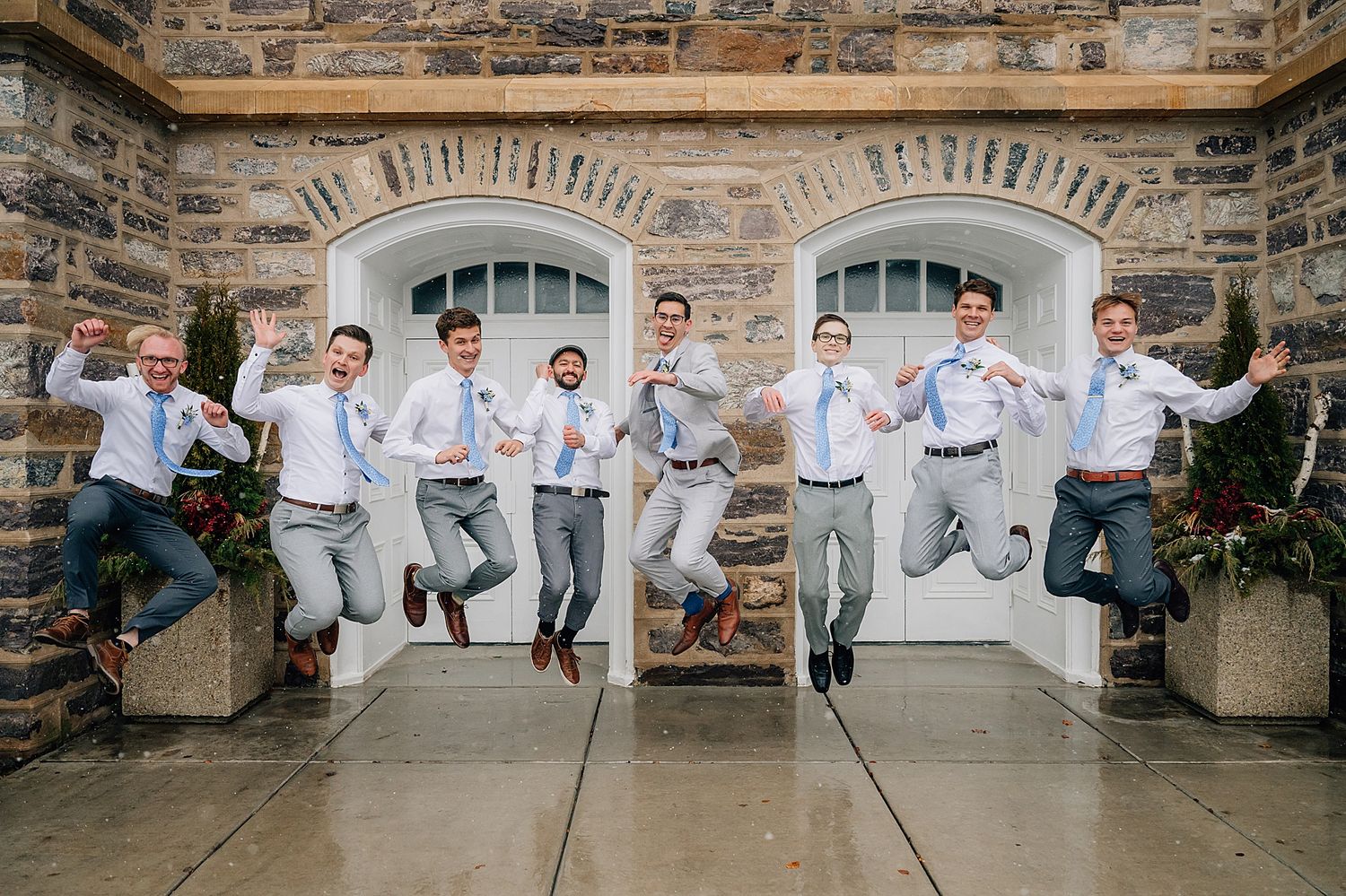 Groomsmen in light gray suits jumping in synchronized pose against stone wall backdrop during wedding photoshoot.