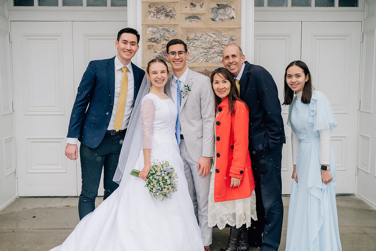 Wedding party in formal attire poses against white garage doors and stone wall on special day.