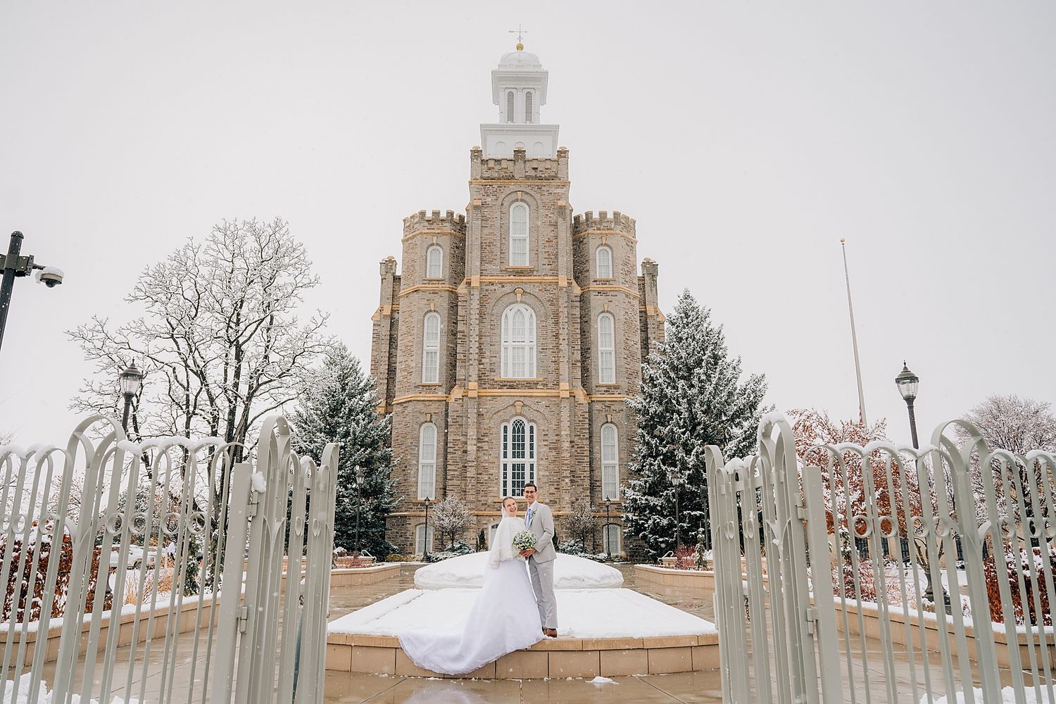 A snow-covered Mormon temple with ornate spires and Victorian Gothic architecture stands behind white iron gates.