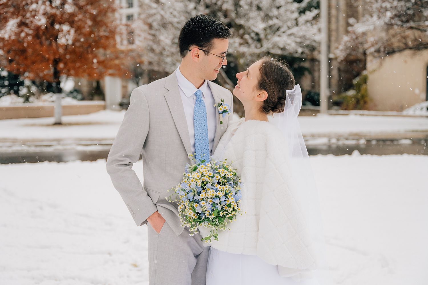 A couple in winter wedding attire share a loving moment in the snow, with the bride holding a blue and white bouquet.