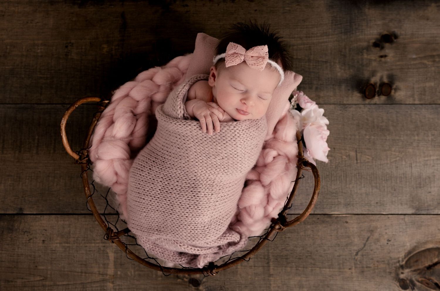 Newborn baby wrapped in soft pink blanket sleeping peacefully in rustic metal basket with pink fabric padding.