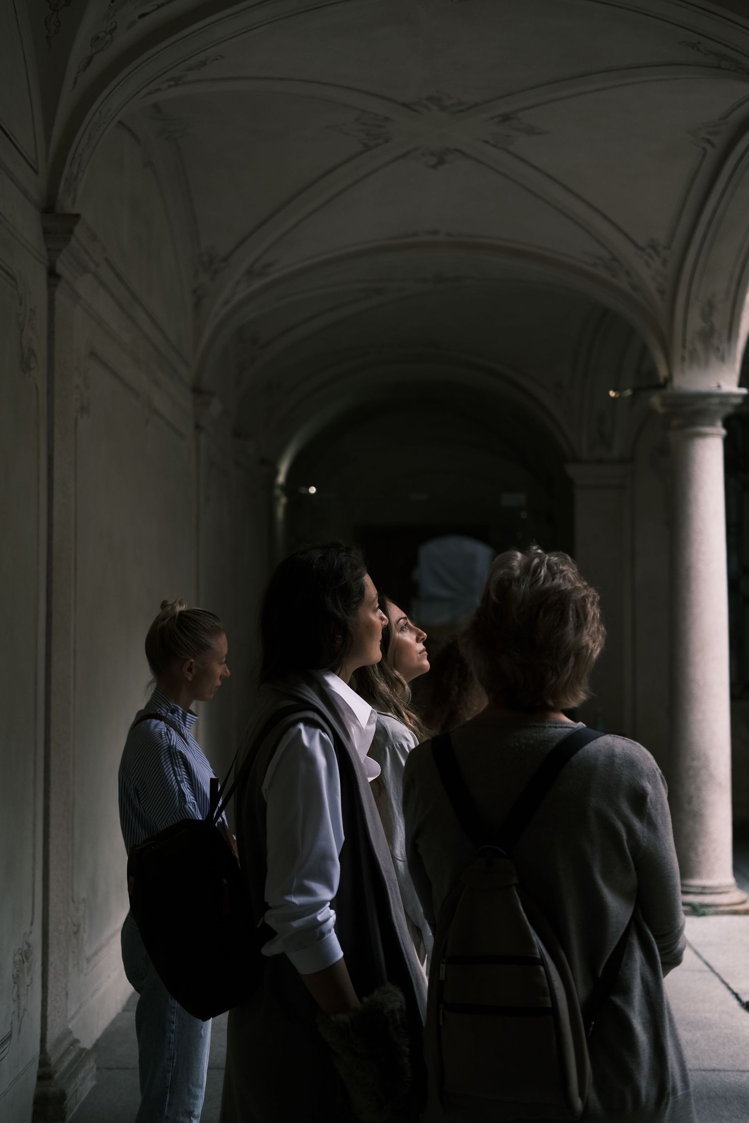 Group of people standing in shadowy arched hallway with classical architecture.