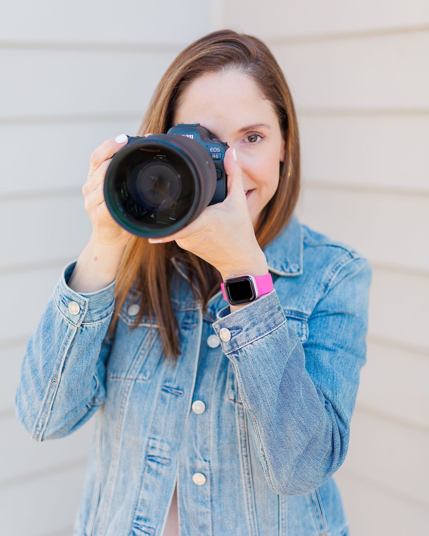 Photographer in denim jacket holds professional camera while wearing smart watch with pink band.