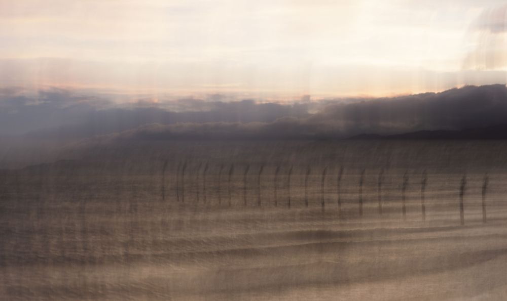 A blurry abstract landscape with dark silhouettes of fence posts or poles against a hazy sunset sky.