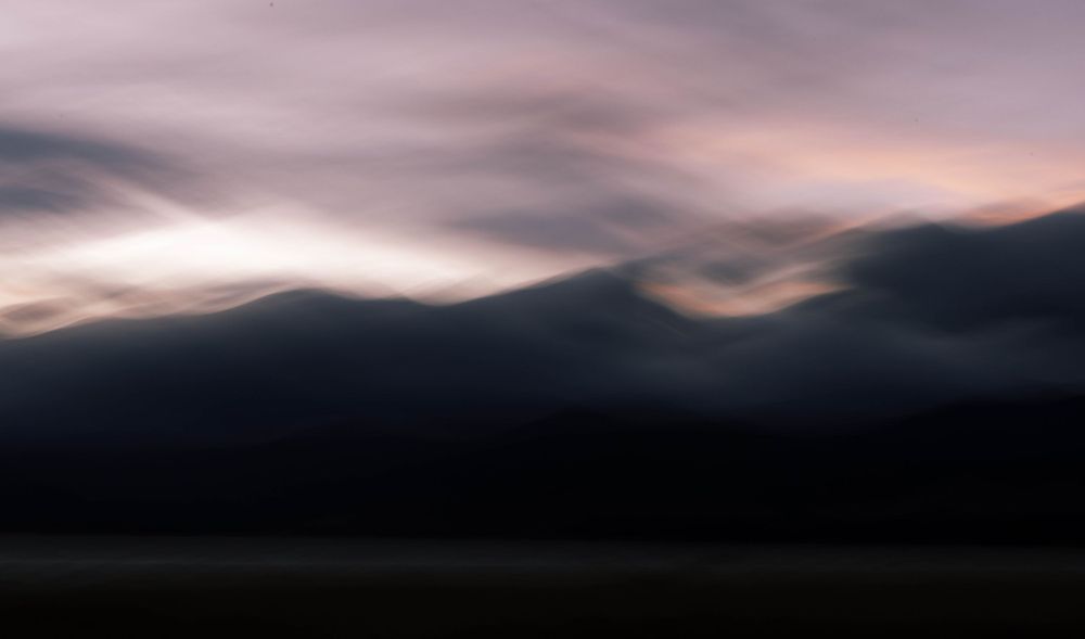 Abstract landscape with dark silhouetted mountains against a moody purple and pink sunset sky with streaking clouds.