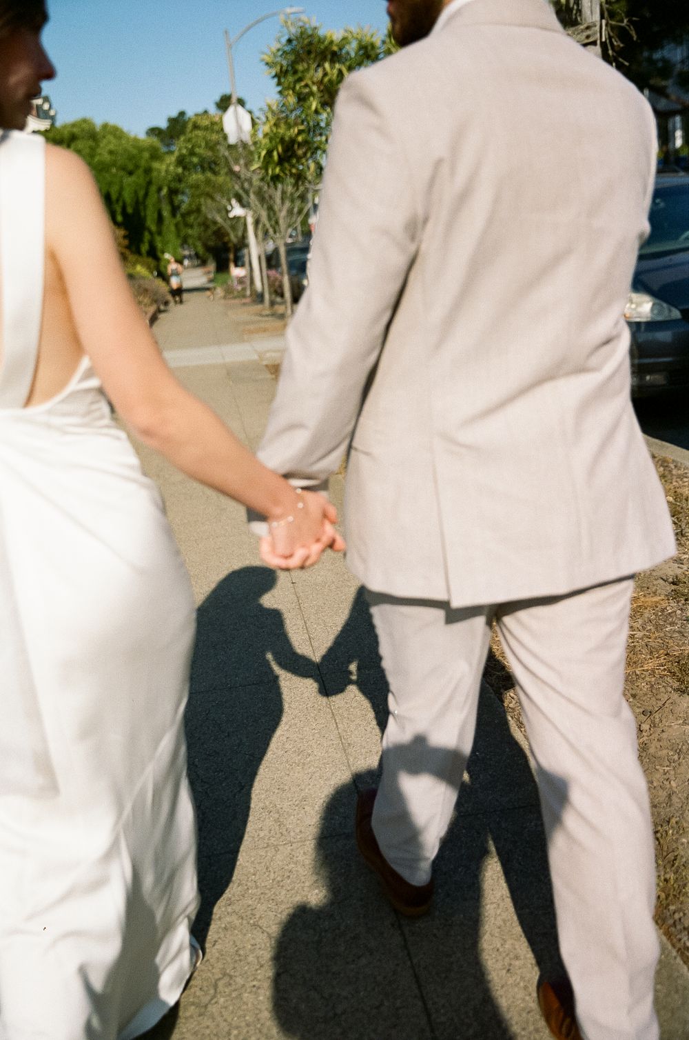 A couple in formal attire holding hands while walking, casting shadows on a sunny sidewalk.