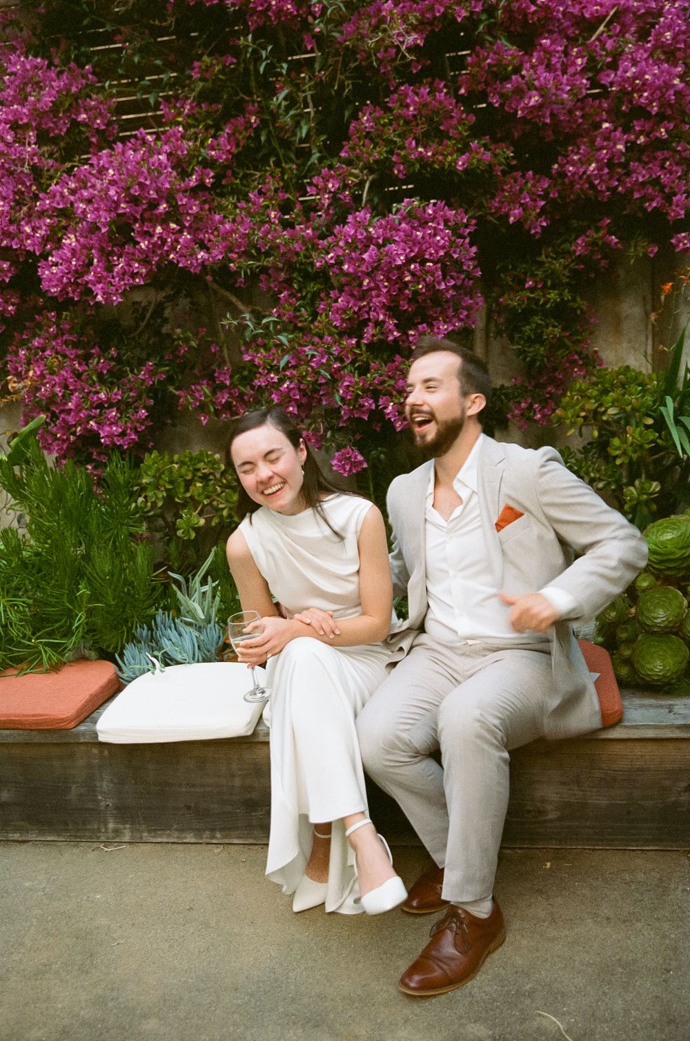 A couple in light colored outfits sits laughing together on a bench beneath vibrant pink bougainvillea flowers.