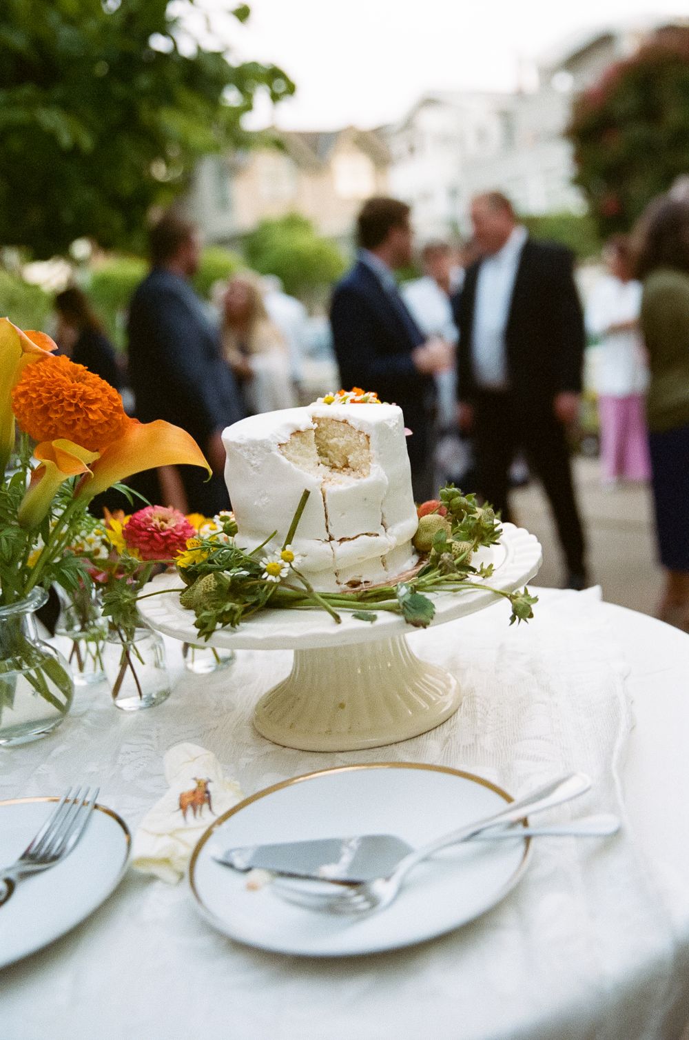 A white wedding cake on a pedestal sits on an elegant table with place settings and orange floral arrangements.