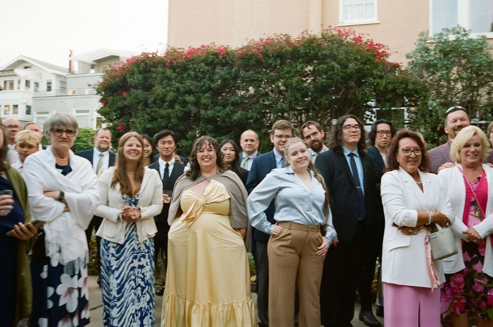 A group of well-dressed guests gather outdoors at a social event with flowering bushes and buildings in the background.