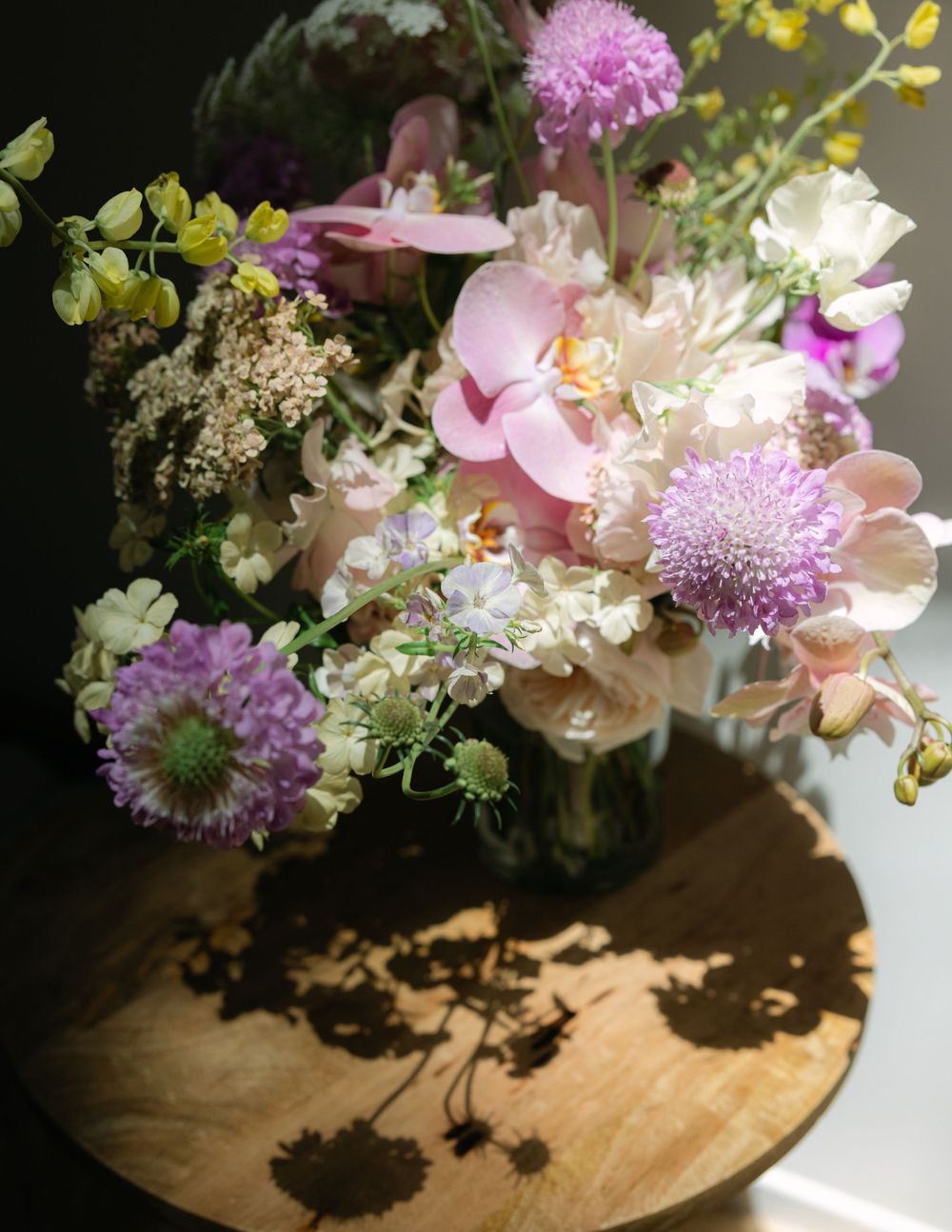 A delicate floral arrangement with pink and purple blooms casts dramatic shadows on a wooden surface in moody lighting.