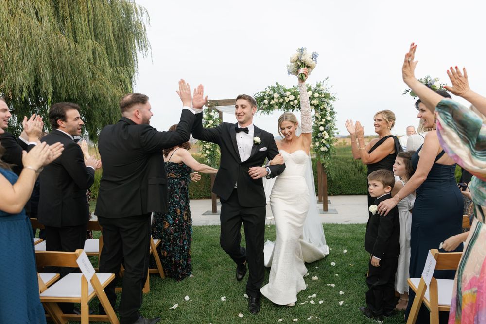 Wedding celebration outdoors with guests cheering and tossing confetti under a willow tree.