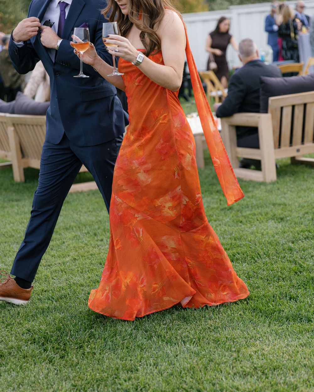 Two guests at an outdoor event with wine glasses, one in a flowing orange dress and another in a navy suit on grass.
