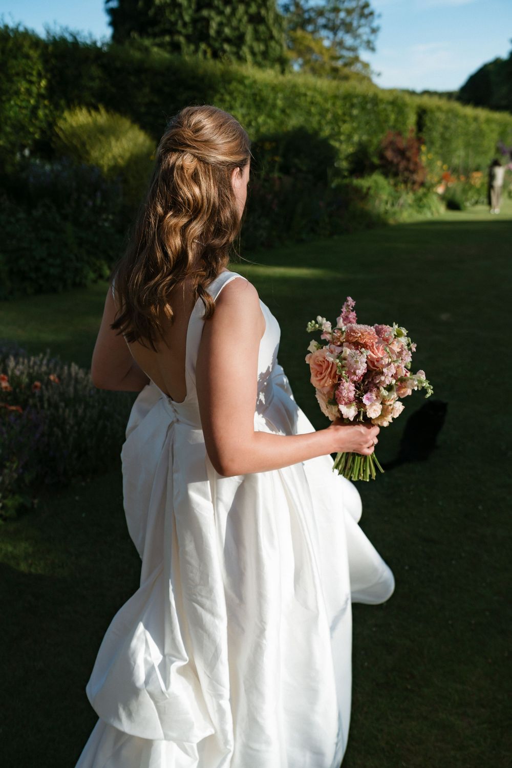 A bride in a flowing white dress holds a pink and peach floral bouquet while walking through a garden setting.
