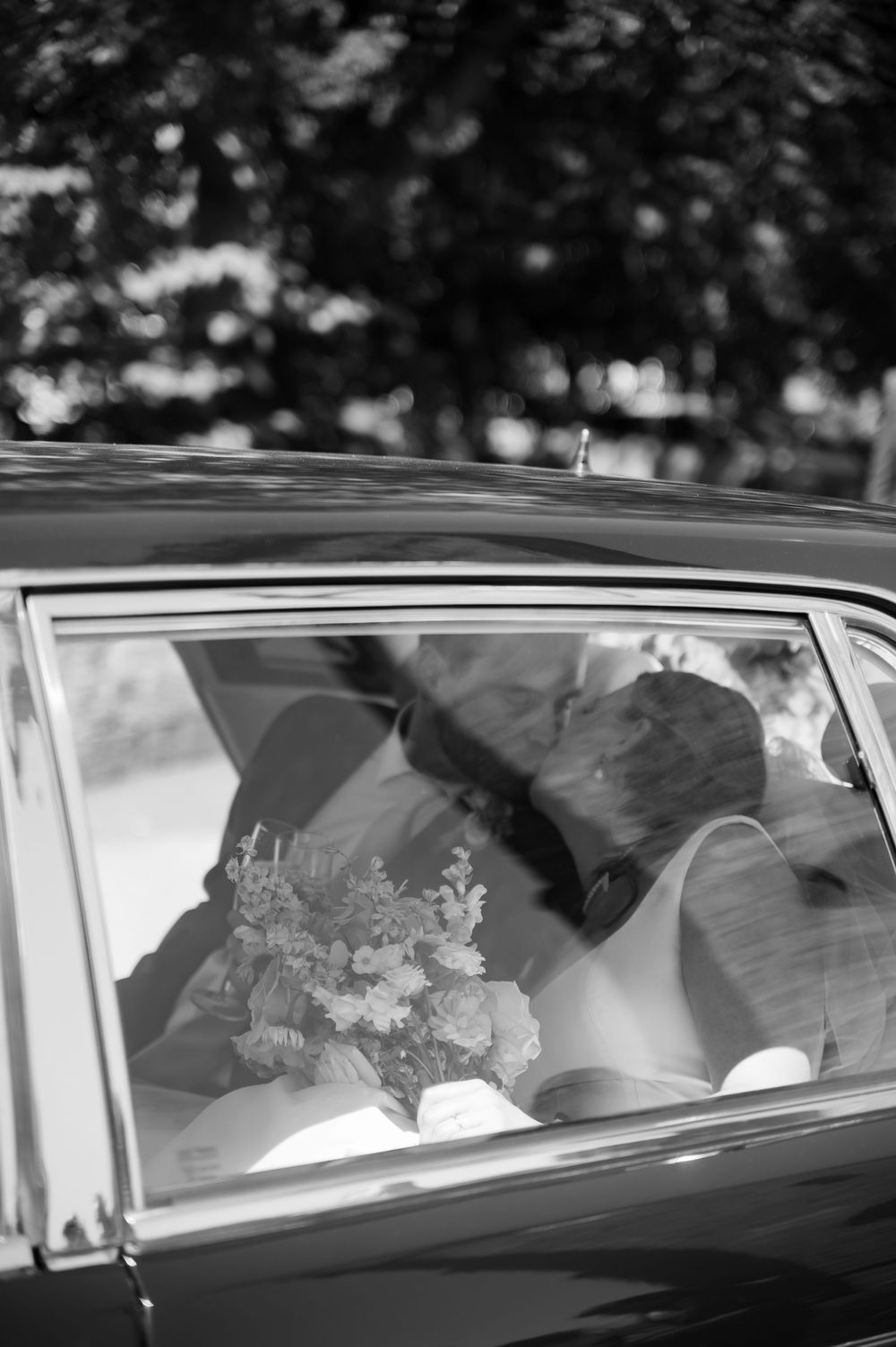 A black and white photograph showing a couple kissing in the back of a vintage car through the window.