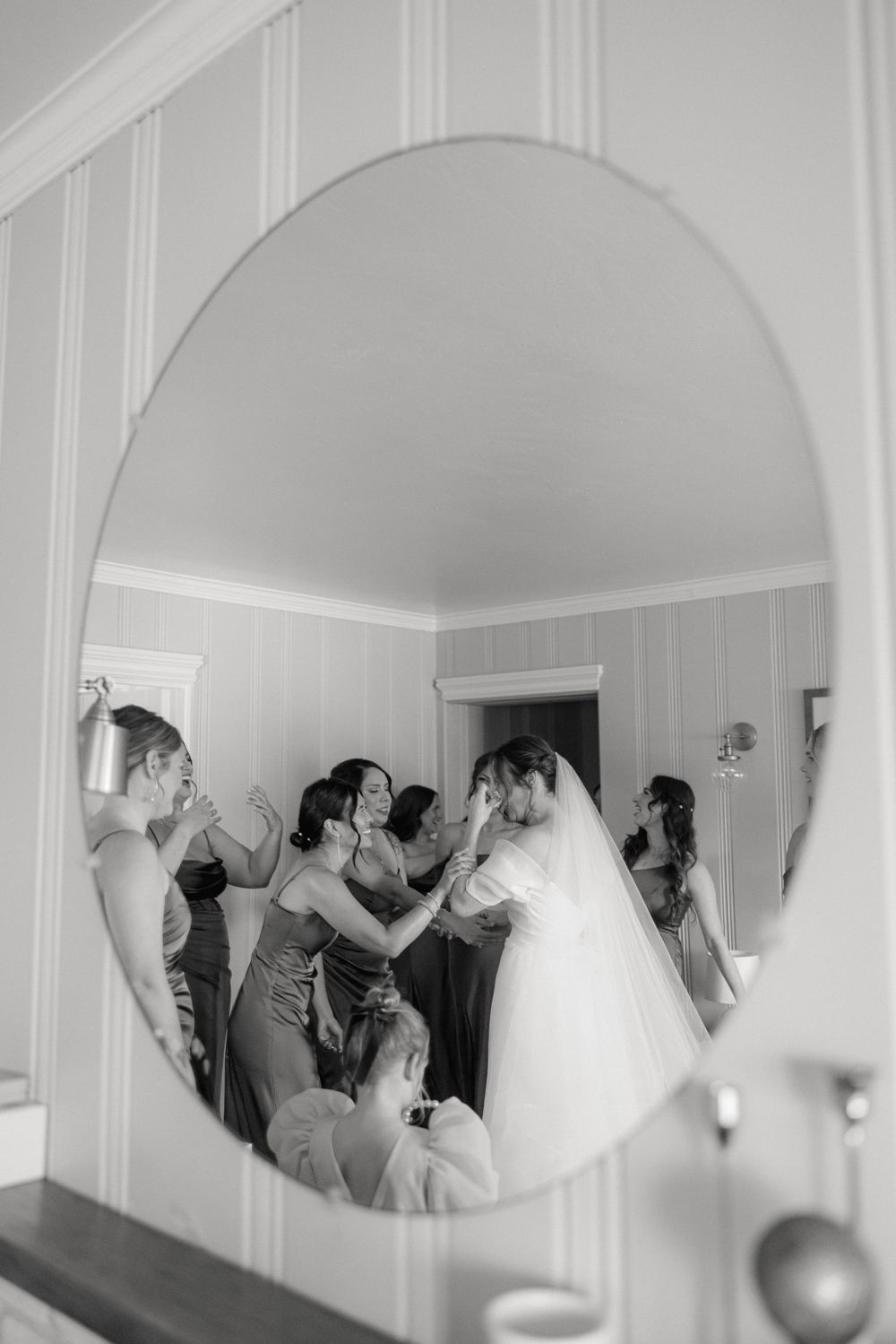 A black and white reflection in an oval mirror shows a bridal party getting ready with bridesmaids helping adjust the wedding dress.