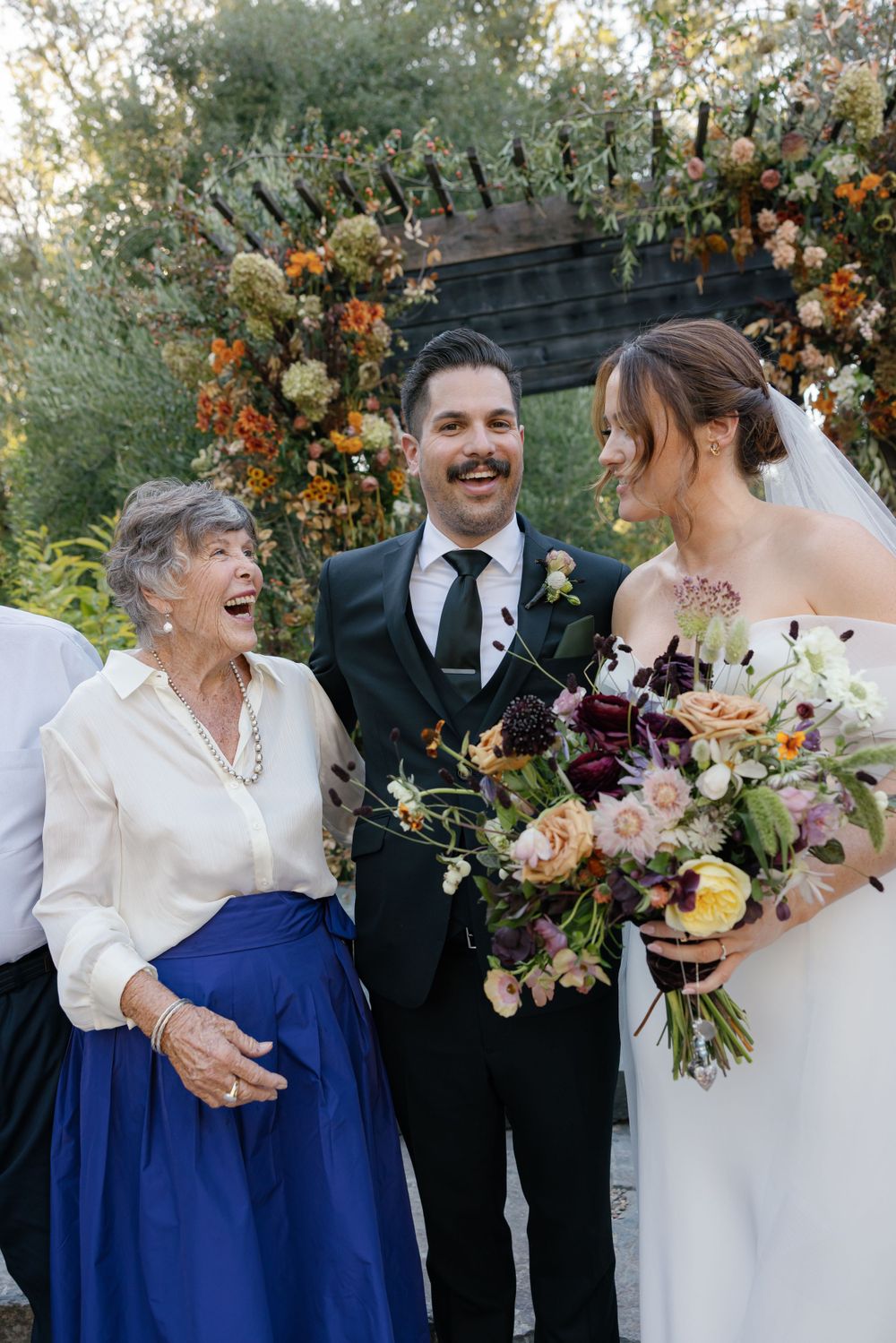 Three wedding attendees share a joyful moment outdoors in front of floral decorations with orange and white blooms.