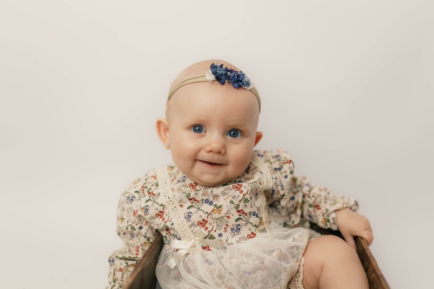Smiling baby in floral dress with blue bow headband poses for portrait.