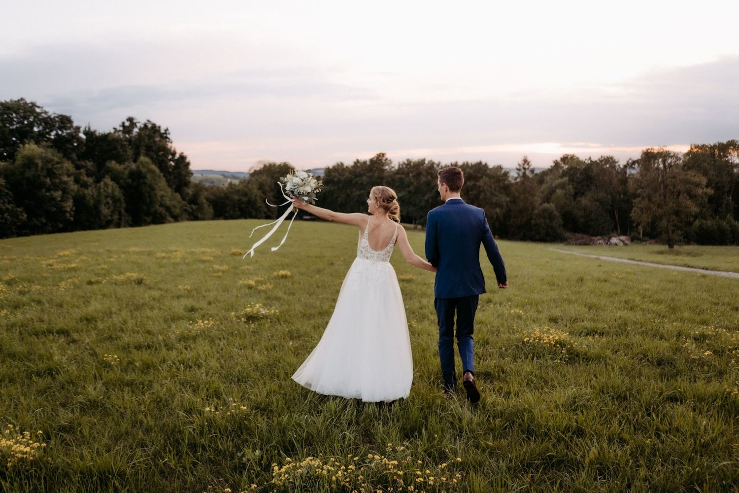 A couple walks hand in hand across a grassy field during their wedding photoshoot.