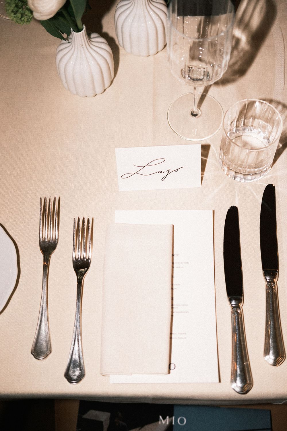 Elegant wedding place setting with calligraphy name card, silver forks, knives, linen napkin, and crystal glasses on a neutral tablecloth.