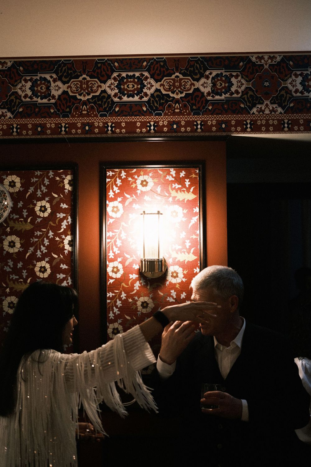 Elegant couple sharing a romantic moment at a dimly lit venue with ornate red patterned walls and candlelight.