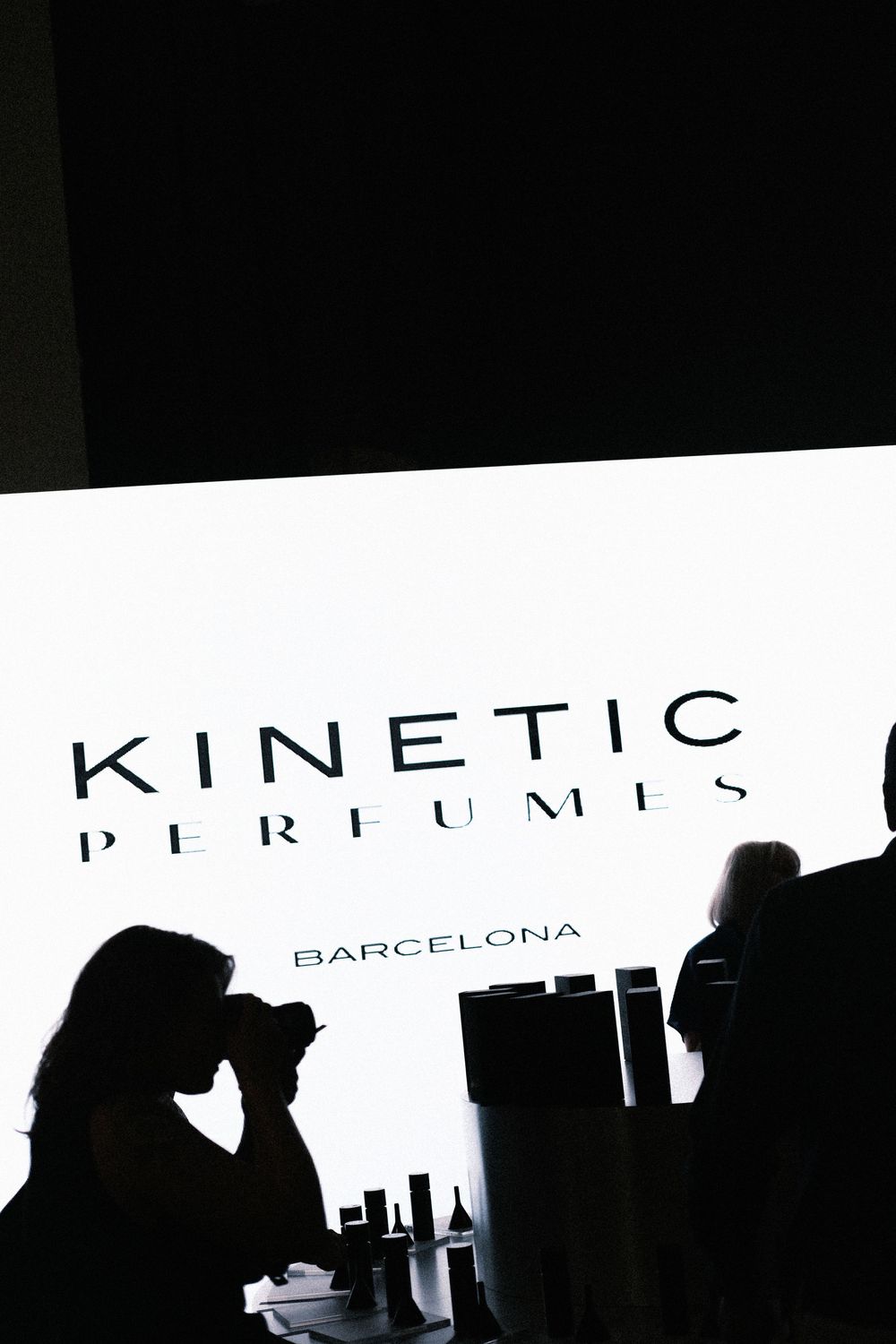 Silhouettes of people in front of a bright Kinetic Perfumes Barcelona illuminated sign display.