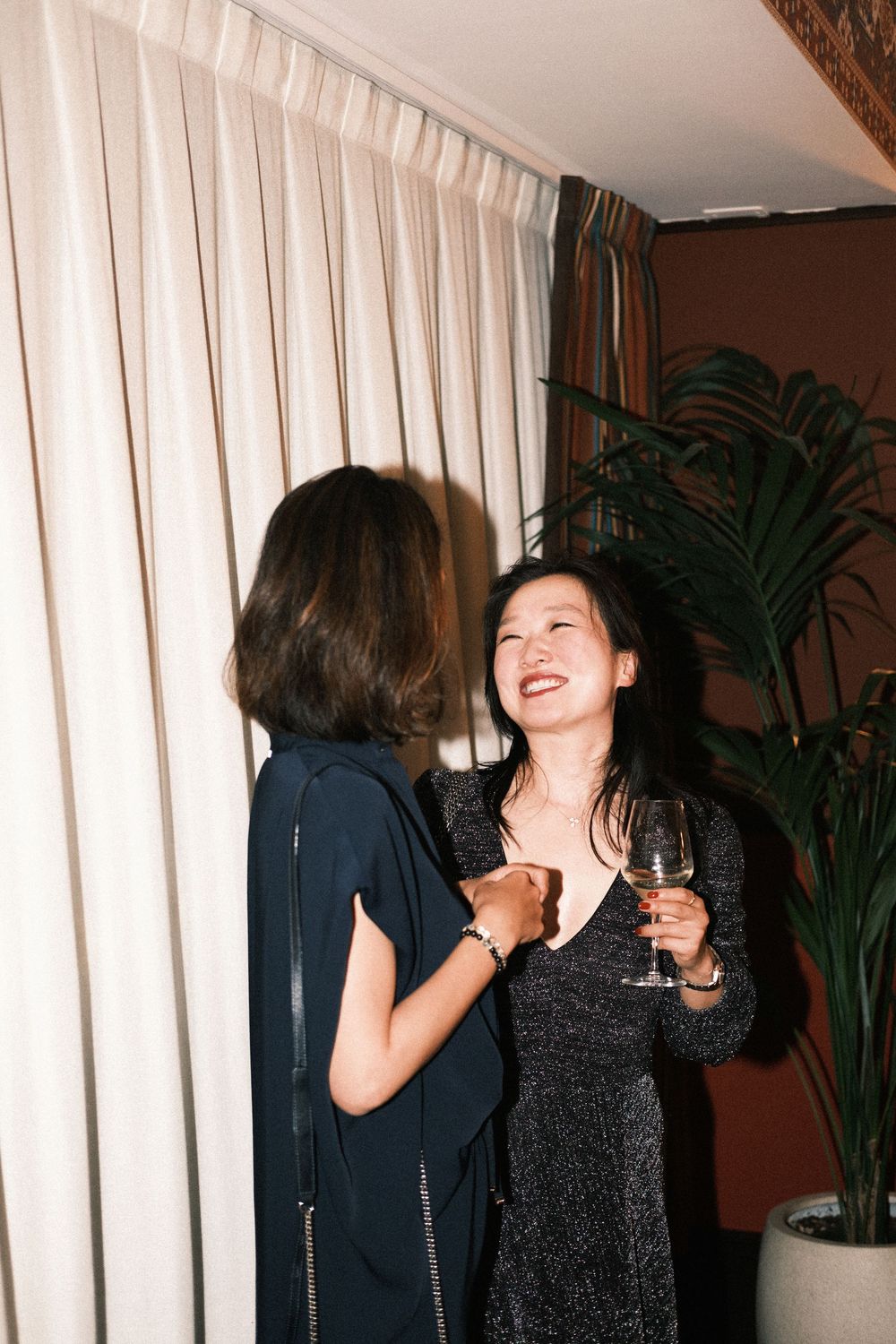 Two women in elegant dresses laughing and socializing at an upscale party, one holding a wine glass.