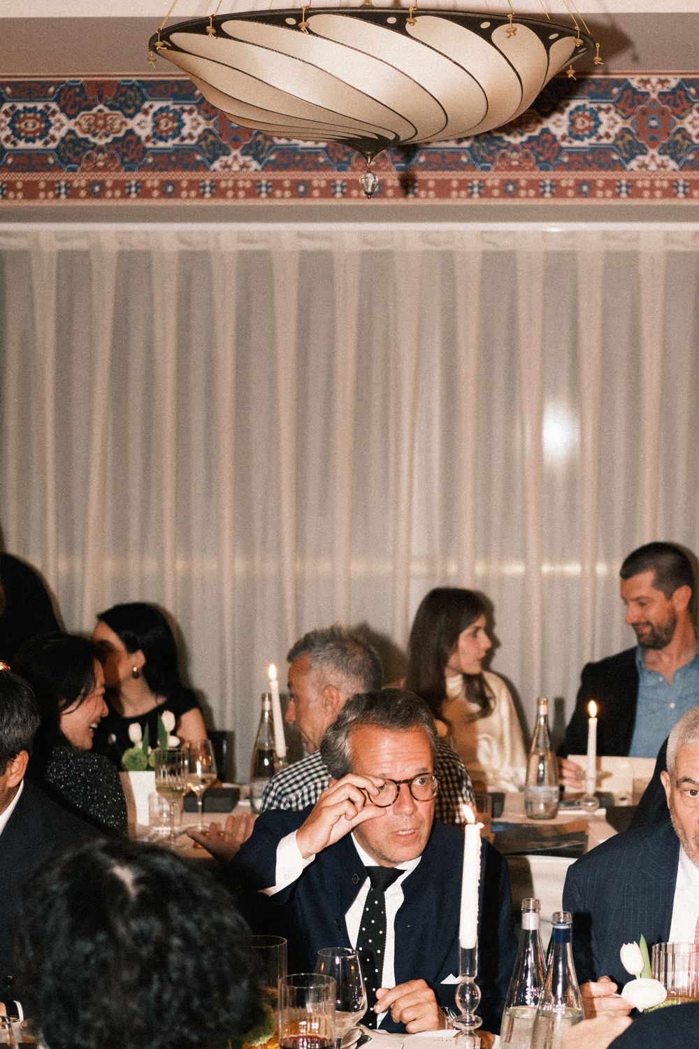 Elegant dinner party guests seated at candlelit tables in a decorated restaurant with sheer curtains.