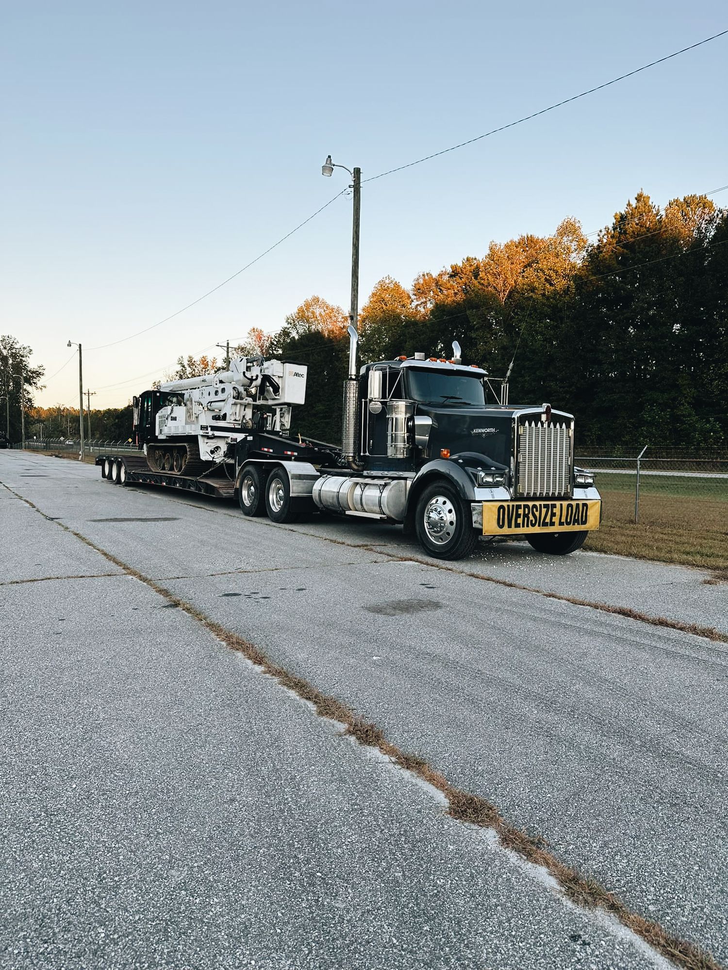 A gray semi truck hauling heavy construction equipment on a flatbed trailer parked on asphalt during golden hour sunset.
