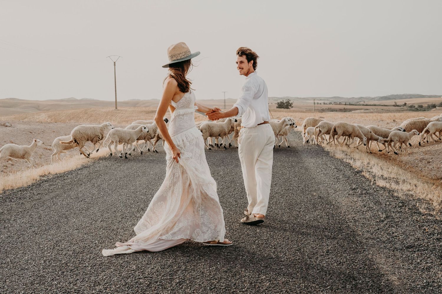 A couple dances on a rural road surrounded by sheep in a dreamy desert landscape during sunset.