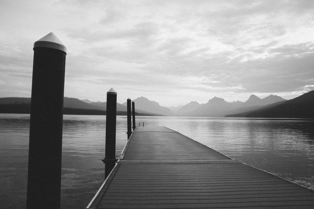 Black and white image of a pier with posts leading to misty mountain views. Glacier National Park.