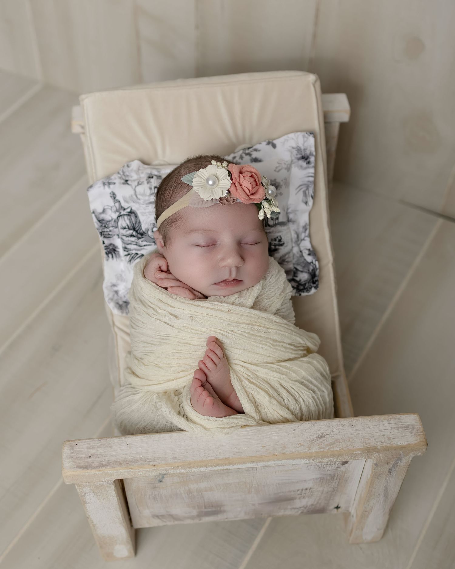 A sleeping newborn swaddled in cream-colored yarn lies on a rustic wooden bed with floral headband and gray floral pillow.