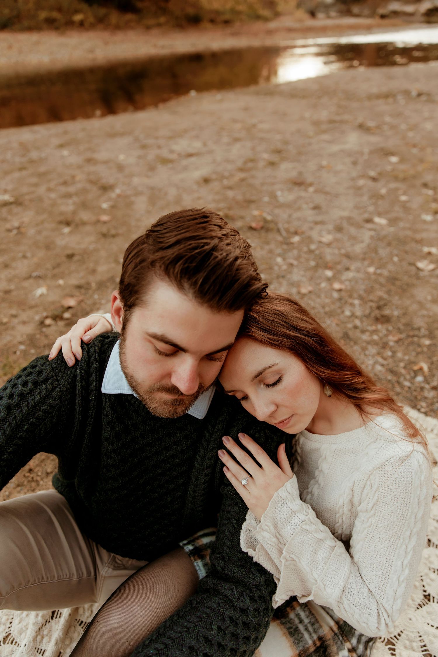 Couple in knit sweaters sharing an intimate moment while sitting by a lake on an autumn day.