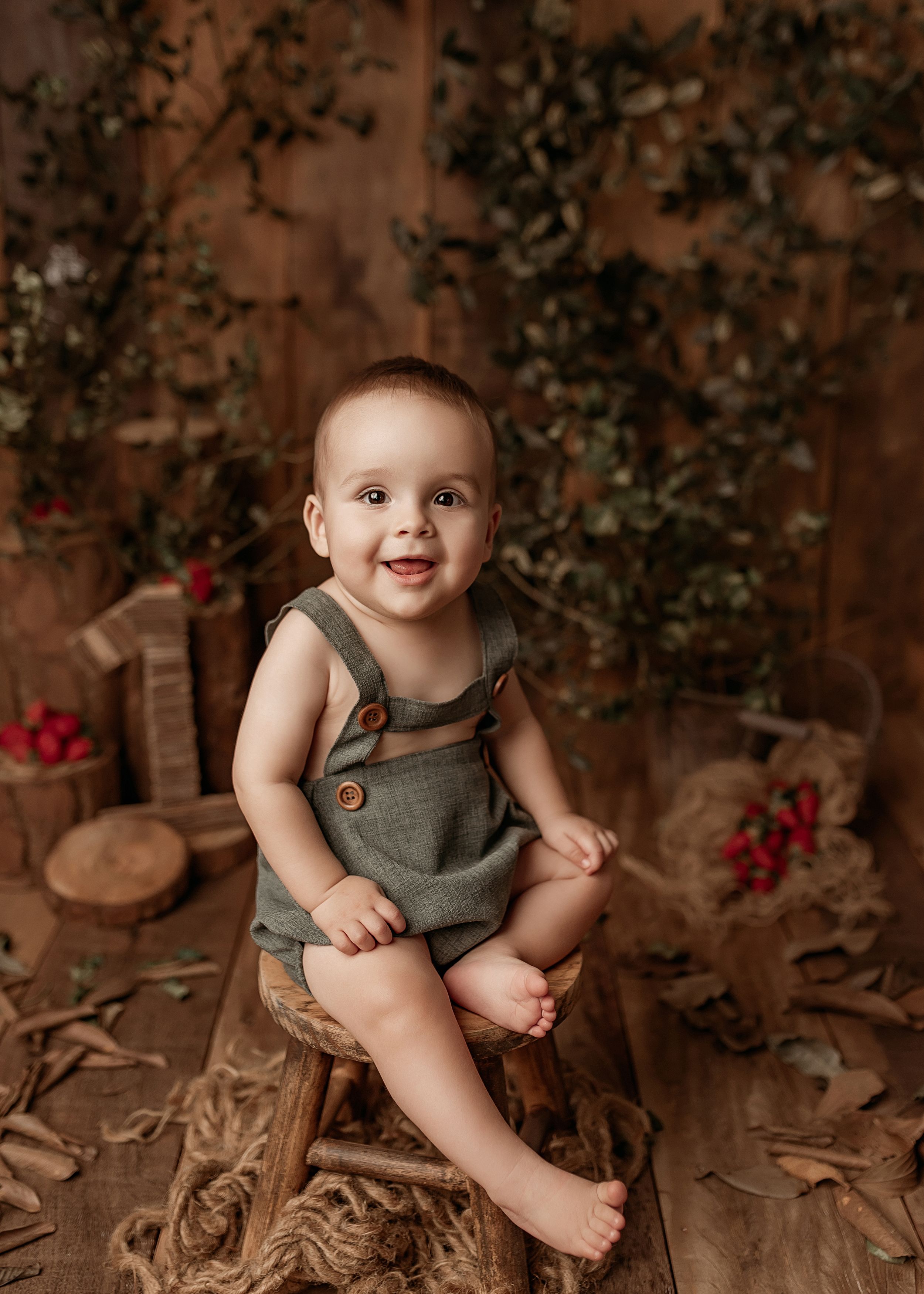 Baby wearing gray overalls smiles while sitting on a rustic wooden chair with red berries in the background.