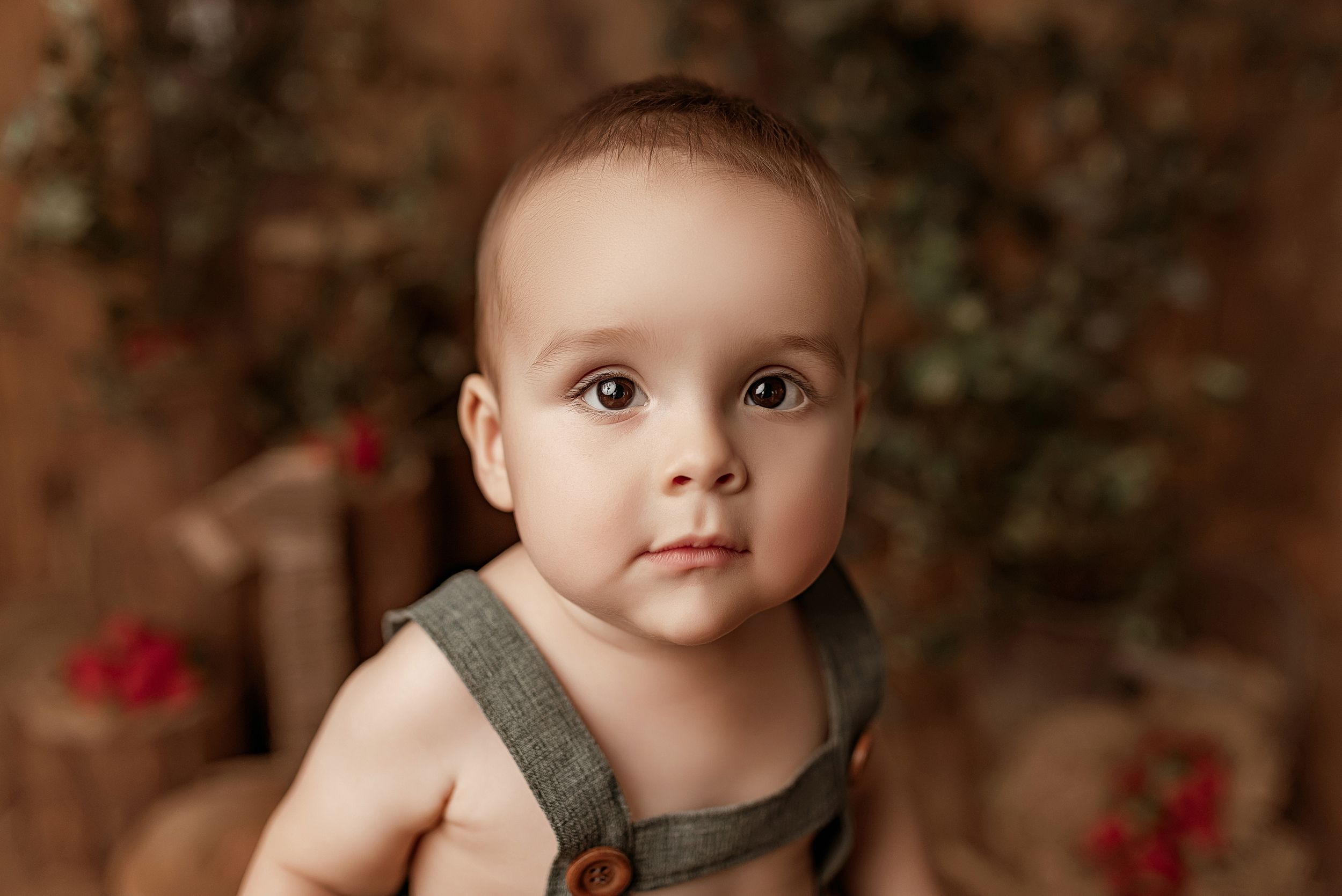 A young child in a sage green tank top poses against a brown backdrop with red flowers in soft lighting.