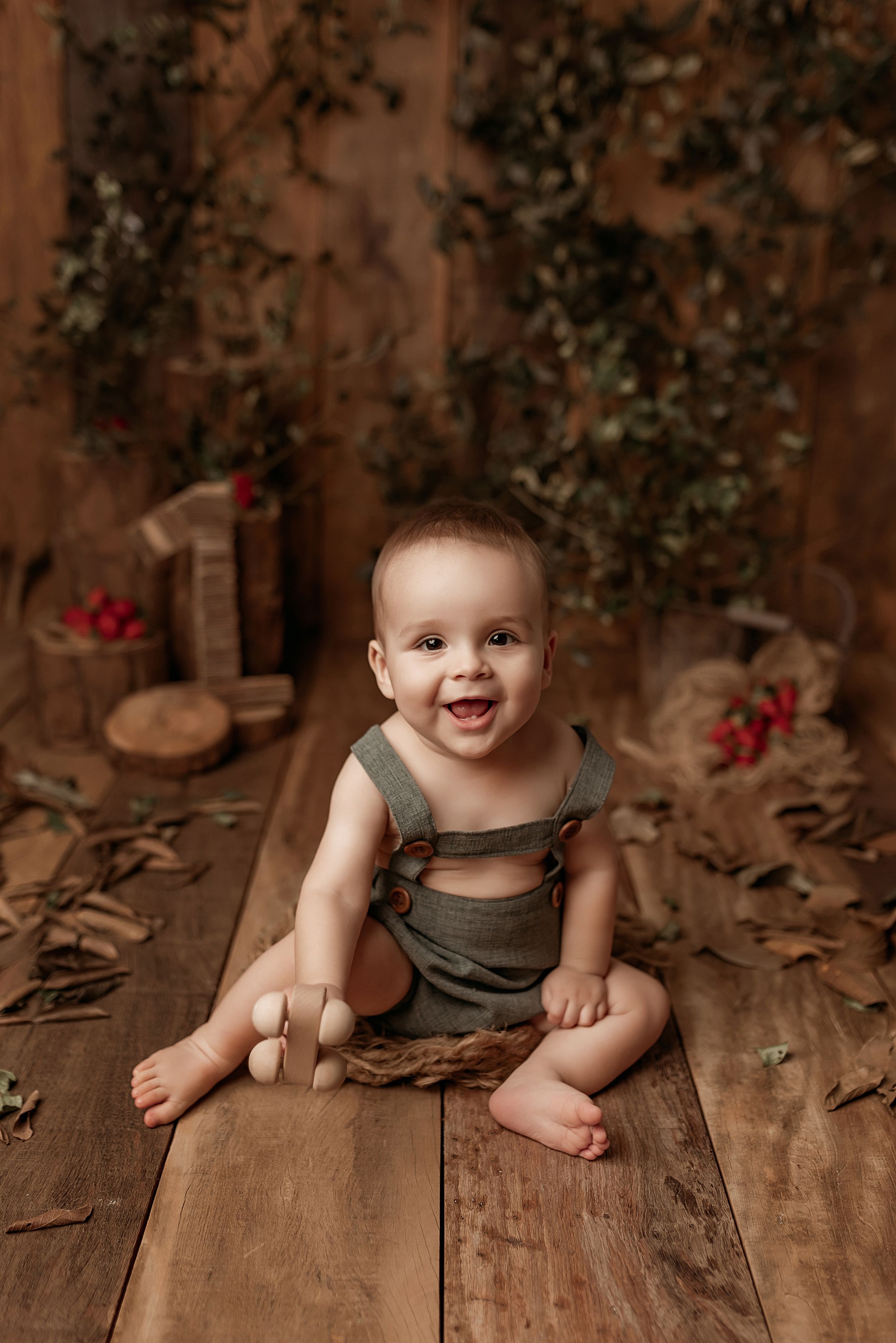 A smiling baby wearing overalls sits on a rustic wooden floor with decorative autumn elements in the background.