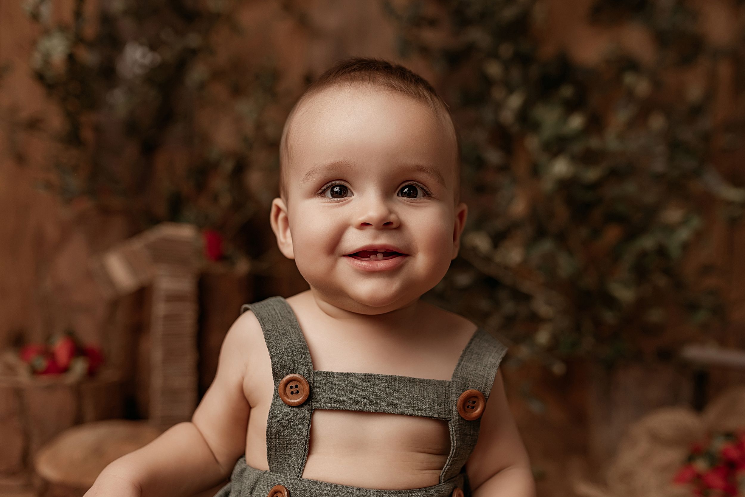 A baby wearing overalls smiles brightly while sitting against a warm brown autumn backdrop.