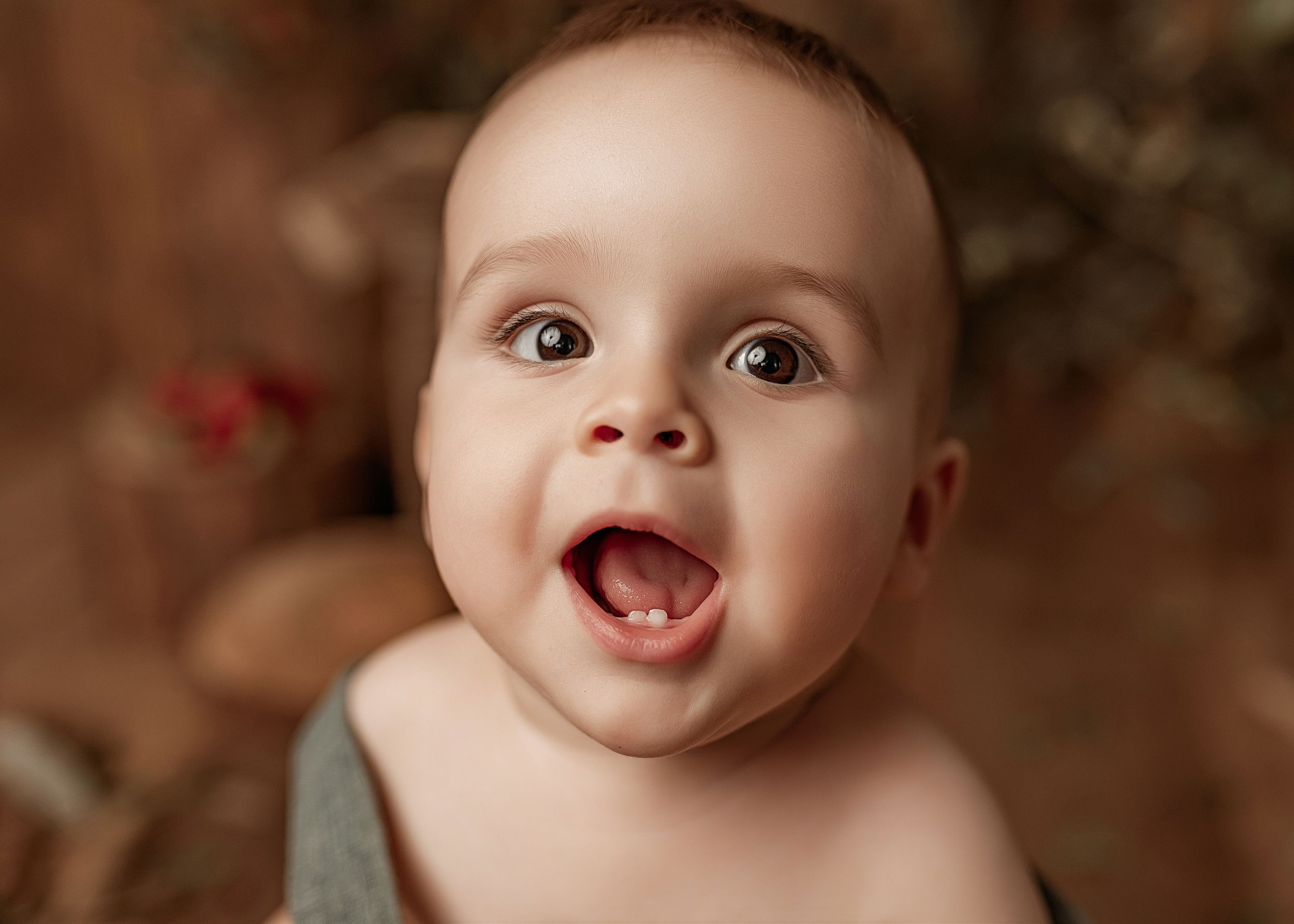 A delighted baby with wide eyes and an open-mouthed smile on a brown background.