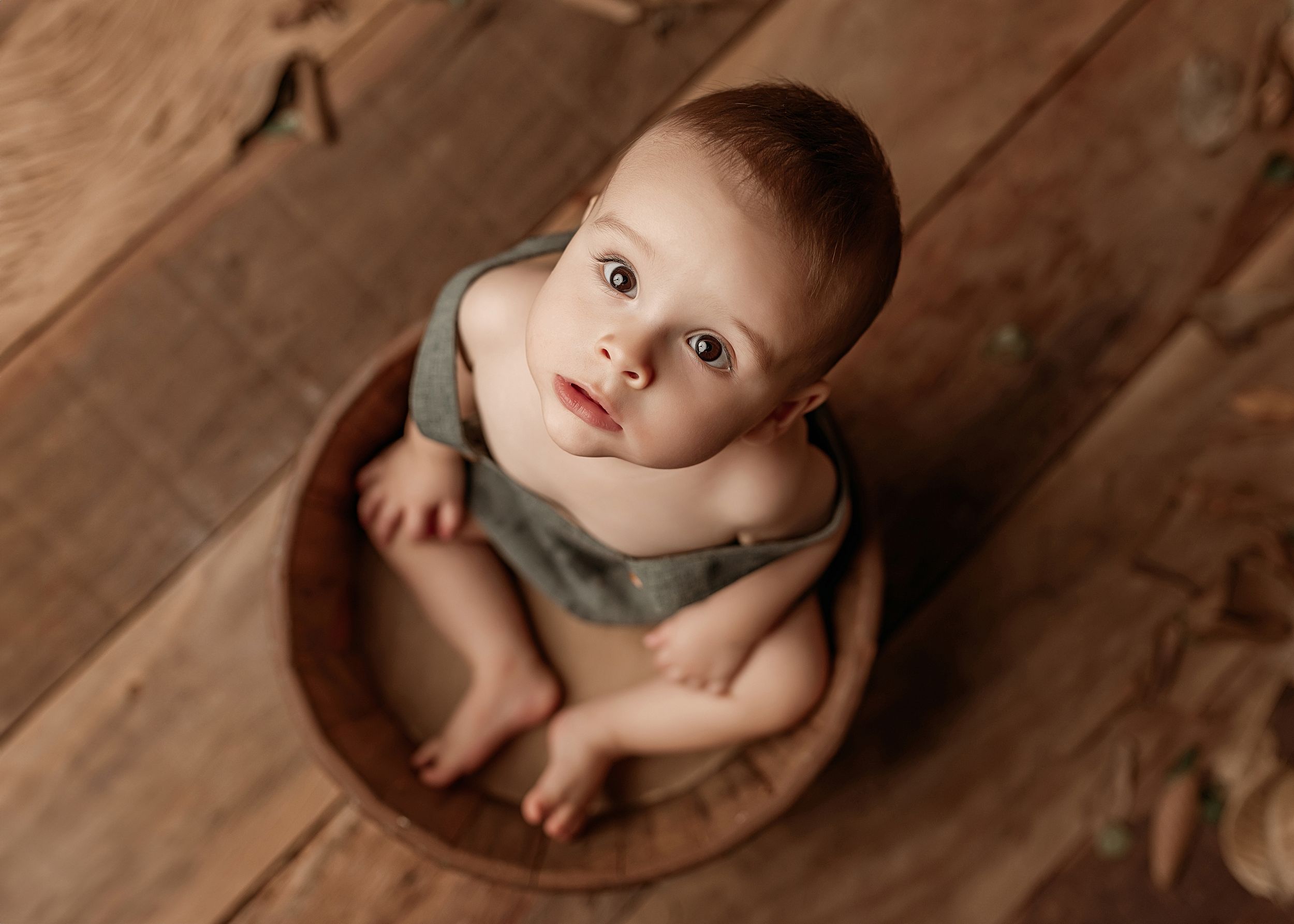 A baby wearing a gray shirt sits inside a wooden bowl on a wooden floor.