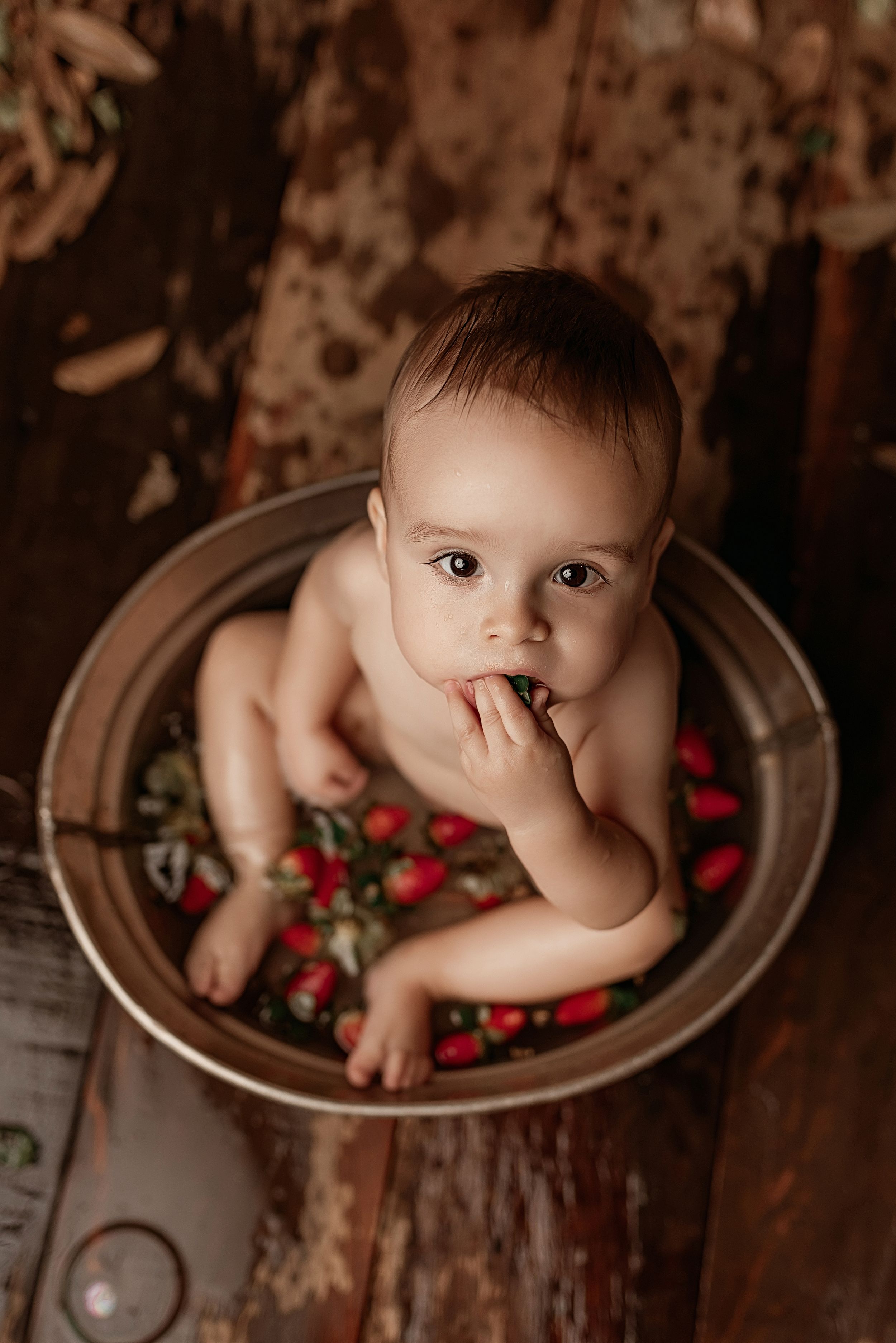 A baby sits in a vintage metal tub filled with red cranberries on a rustic wooden floor for a holiday photo shoot.