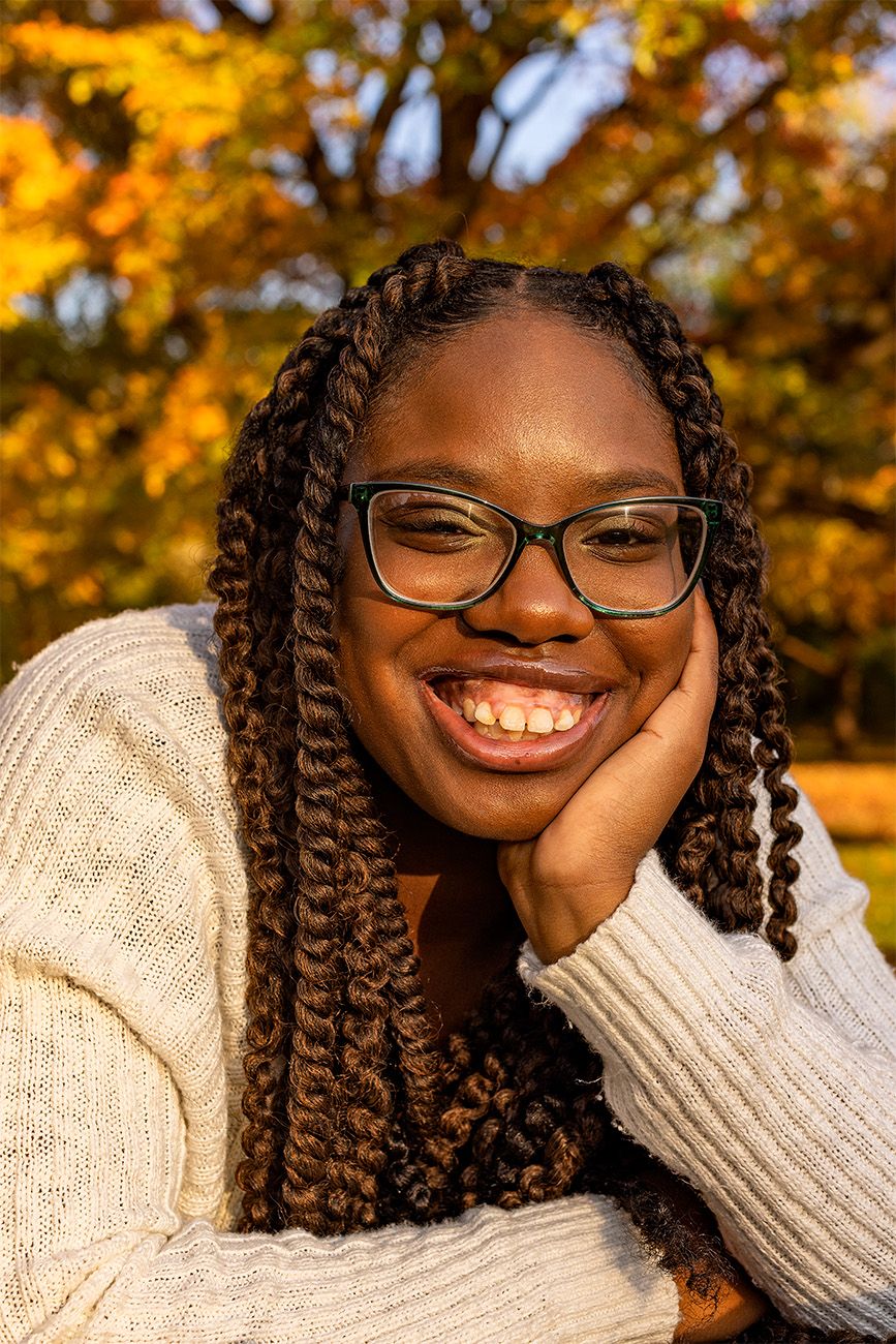 Portrait of a person in a white sweater smiling against fall foliage.