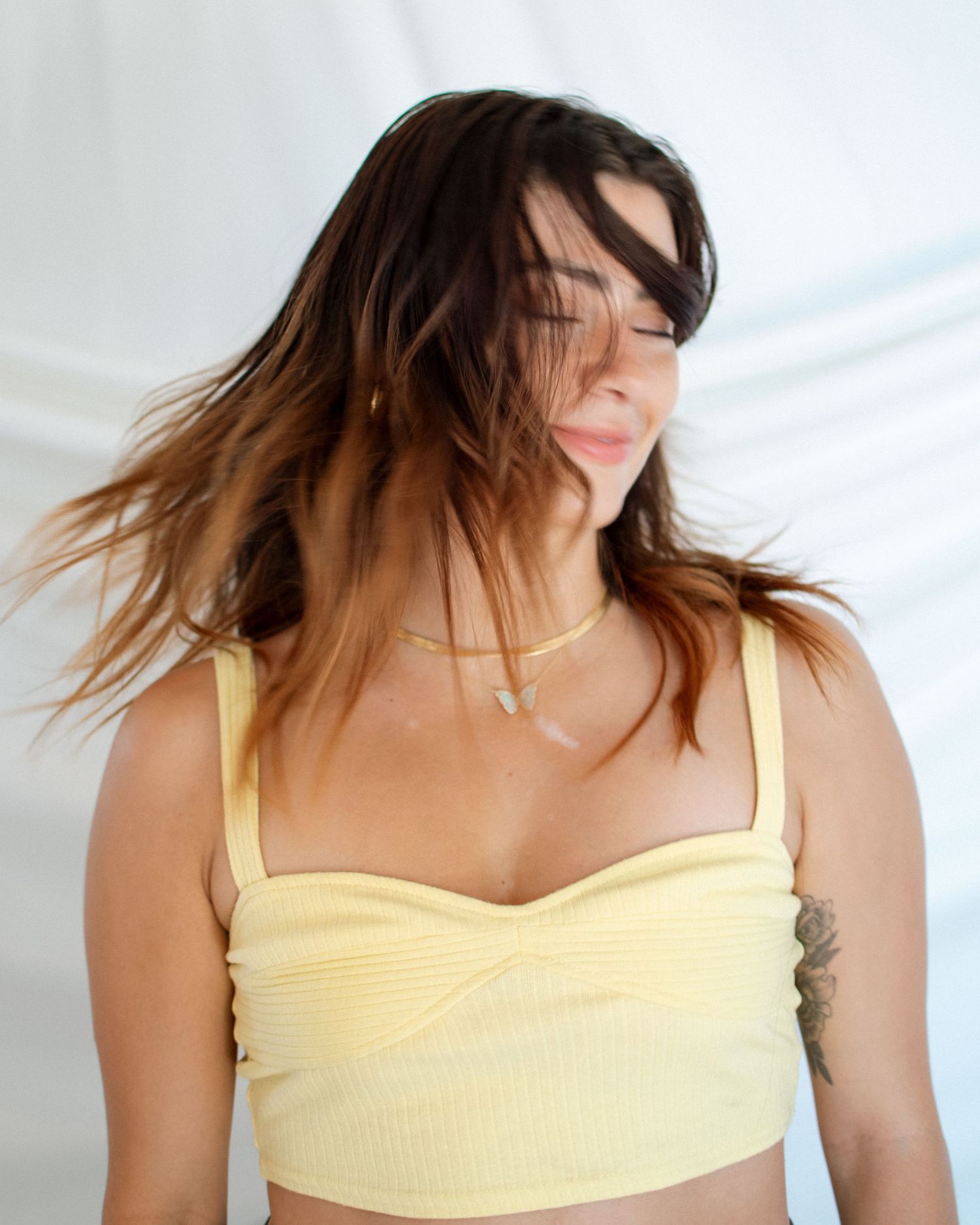 Person with flowing dark hair wearing a pale yellow crop top poses against a white background in natural light.