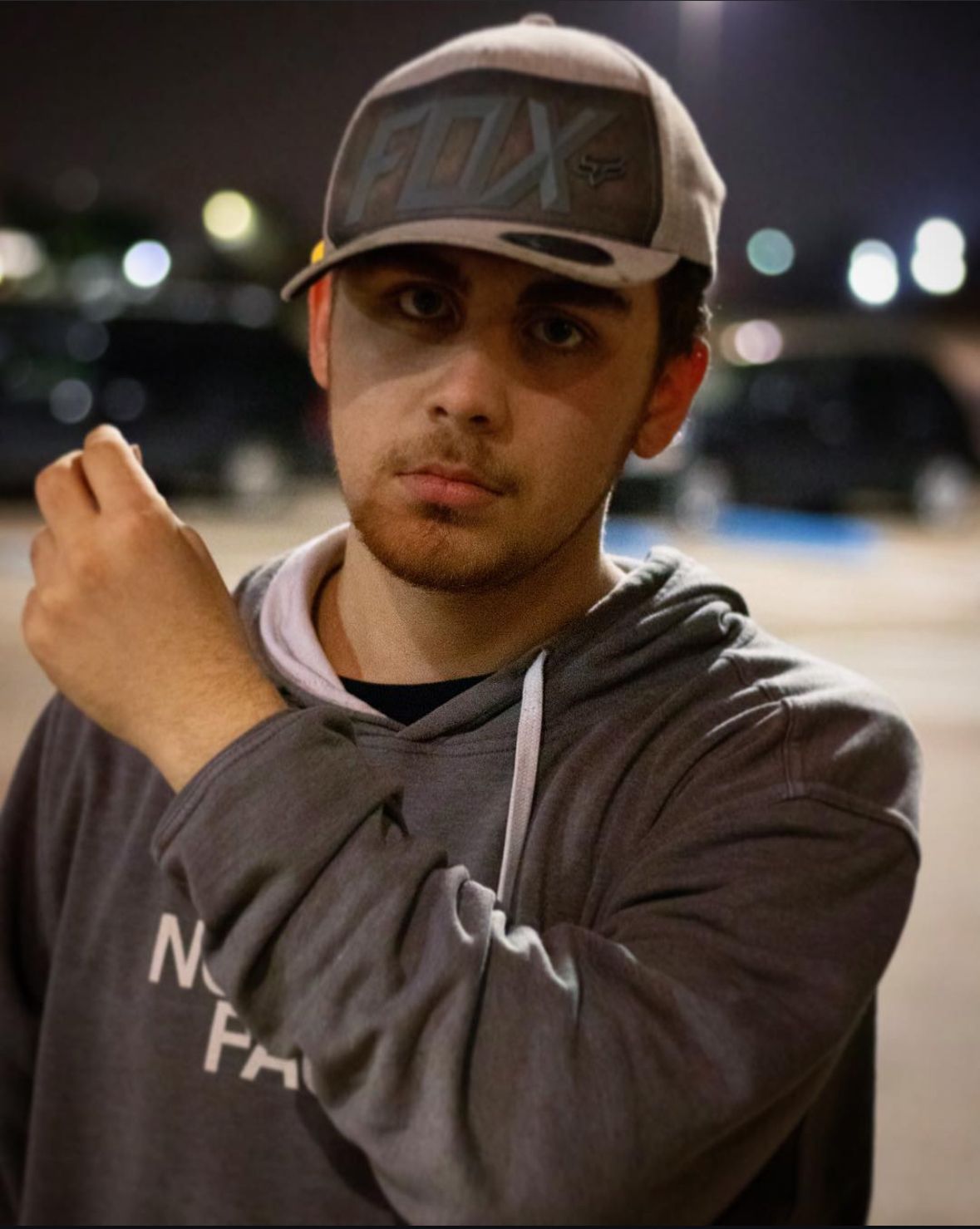 A man poses at night wearing a baseball cap and gray hoodie with blurred lights visible in the background.