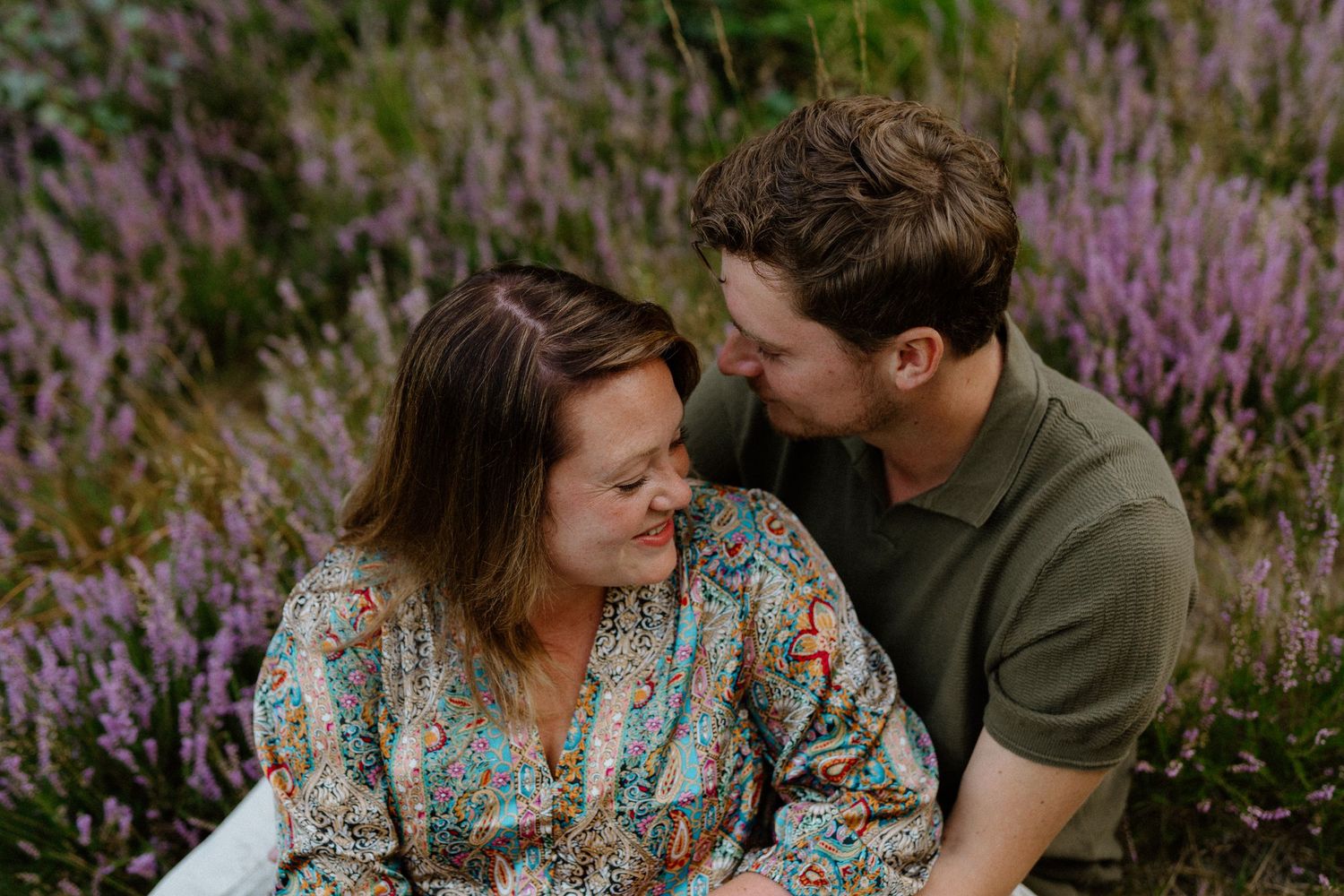 A couple shares a tender moment while sitting among blooming purple wildflowers in a natural outdoor setting.