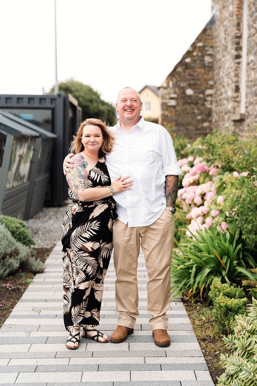 Couple in casual attire smiling together on stone paved walkway with garden background.