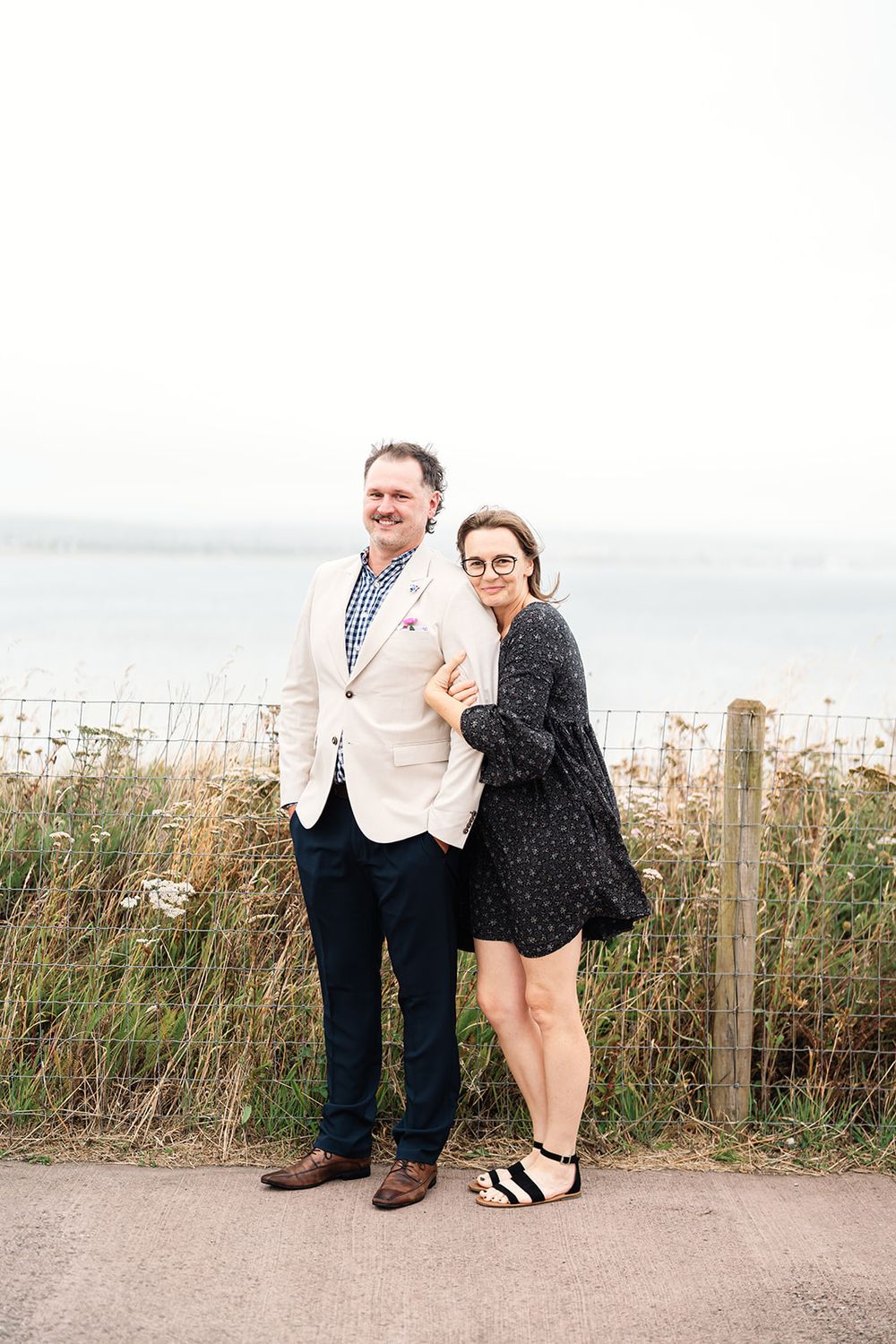 Couple posing together on coastal path with ocean and wild grasses in background.