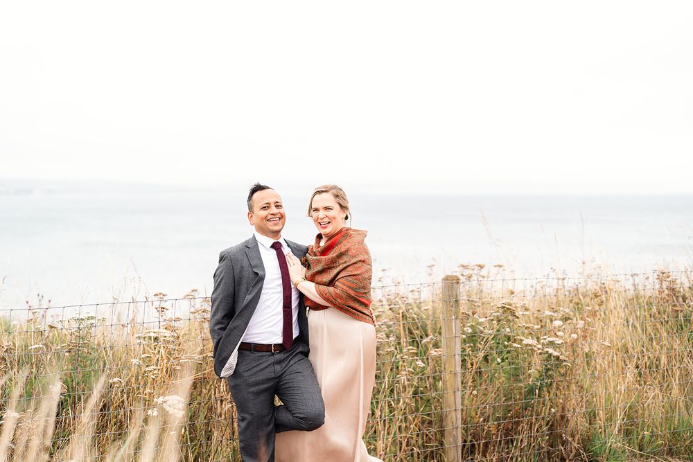 Couple embracing on coastal clifftop path with wild grasses and ocean view.