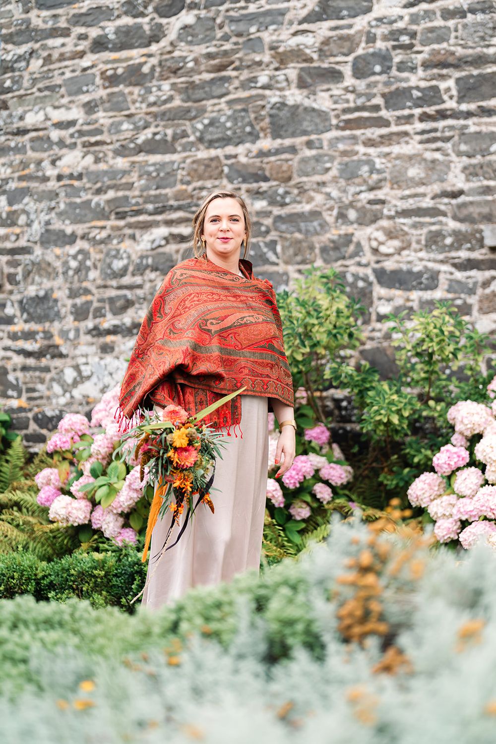 Person in red shawl standing among garden flowers against stone wall background.